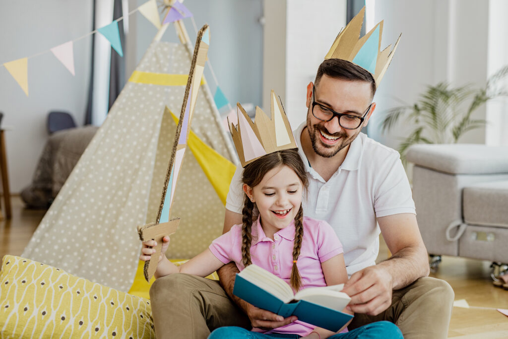 A photo of a young girl and her father, reading a book together wearing paper crowns. The young girl is also wielding a cardboard sword